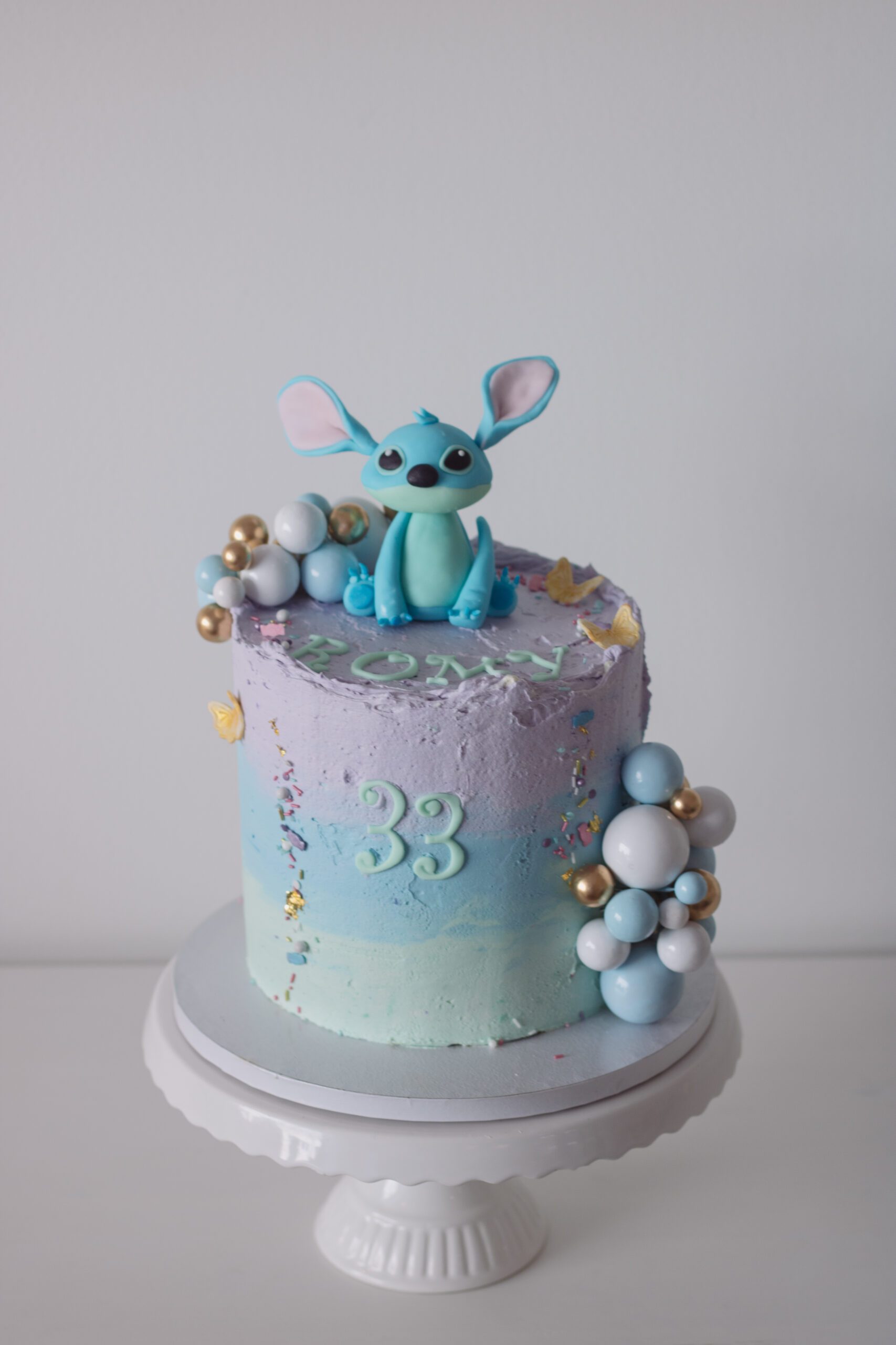 einstöckige Geburtstagstorte mit Farbverlauf von mint zu Lila mit goldenen, weißen und blauen Kugeln und Stitch Figur